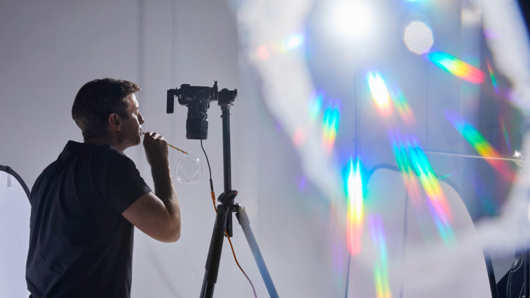 Behind the scenes at a creative photography job shoot, featuring Matt Reed blowing bubbles, camera equipment, and rainbow coloured light reflections.
