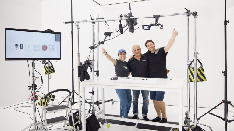 This type of B2B project illustrates "what is commercial photography?". Inside Sum Effect Studio, standing amid the photography and filming equipment at the end of a studio filming session we see the jubilant team and a business owner at the end of a multi-day project Standing and facing the camera with thumbs up, from the left is a female, and then two males. The male in the middle is the business owner, who commissioned our team to make product videos.